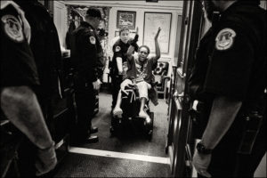 ADAPTer in wheelchair being pushed out of an office by Capitol police officer