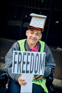 ADAPTer in wheelchair, wearing a house shaped cardboard hat, holding a “Freedom” sign.